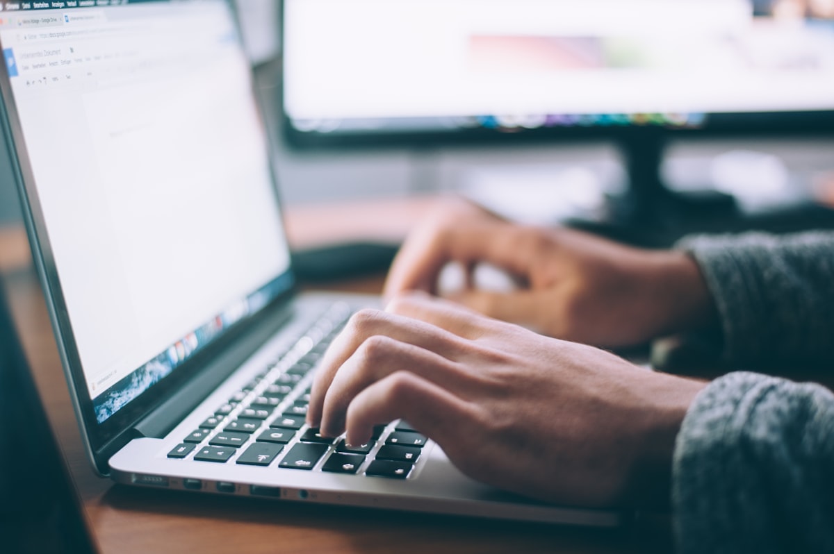 Person typing on a laptop with a second monitor showing code in the background